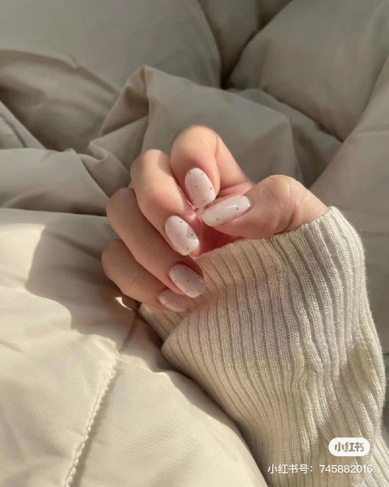 Bàn tay với móng tay sơn màu trắng đốm, áo len màu be, đặt trên chăn mềm mại màu sáng.