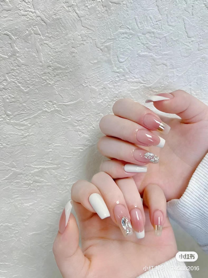Hai bàn tay với móng tay được trang trí màu hồng nhạt, trắng và đính đá, trên nền tường trắng.