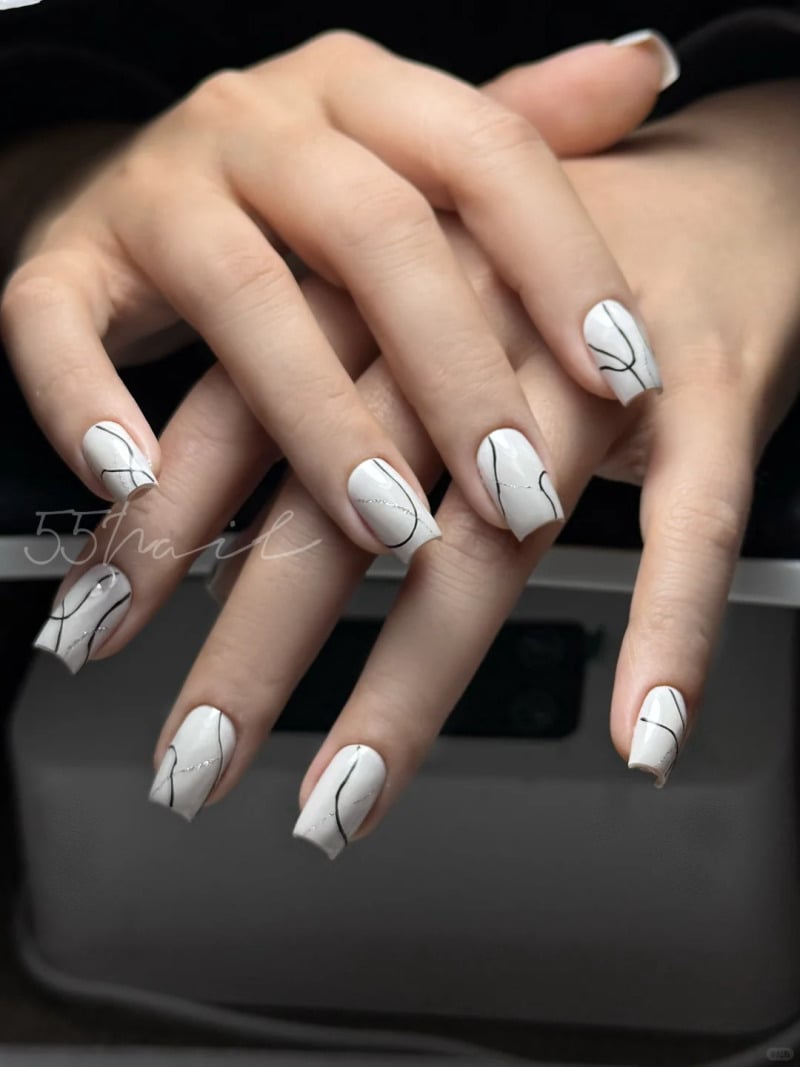 Bàn tay với móng tay sơn màu trắng, trang trí hoa văn đường kẻ đen nghệ thuật, đặt trên nền tối.