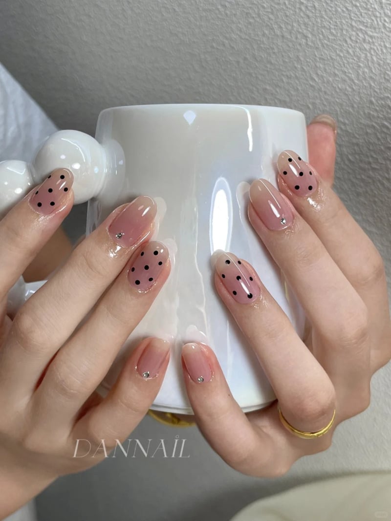 Hai bàn tay với móng tay sơn màu nude và chấm bi đen, cầm một chiếc cốc màu trắng.