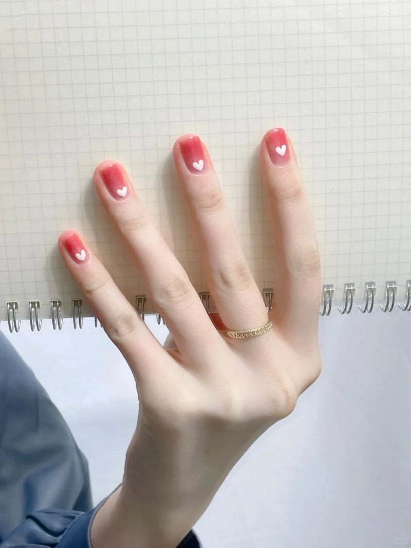 Bàn tay với móng tay sơn đỏ, họa tiết trái tim trắng, đặt trên cuốn sổ lò xo có bìa kẻ ô vuông.