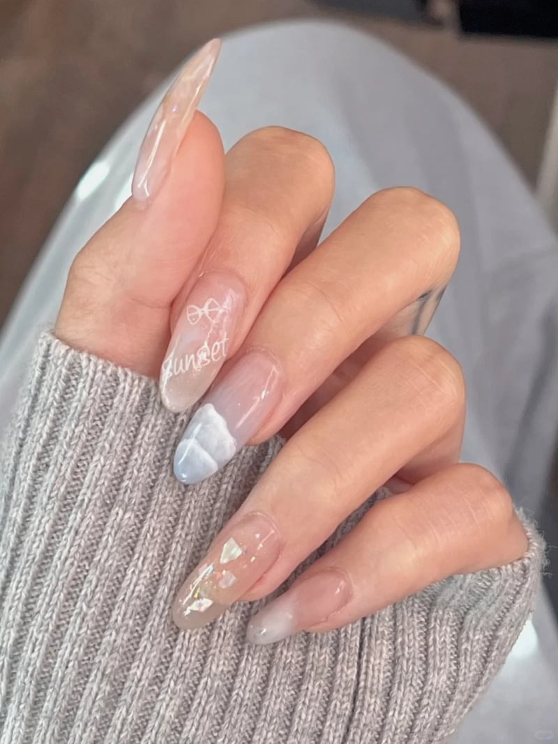 Bàn tay với móng tay nghệ thuật màu pastel, có họa tiết và chữ, trên nền vải áo len xám.