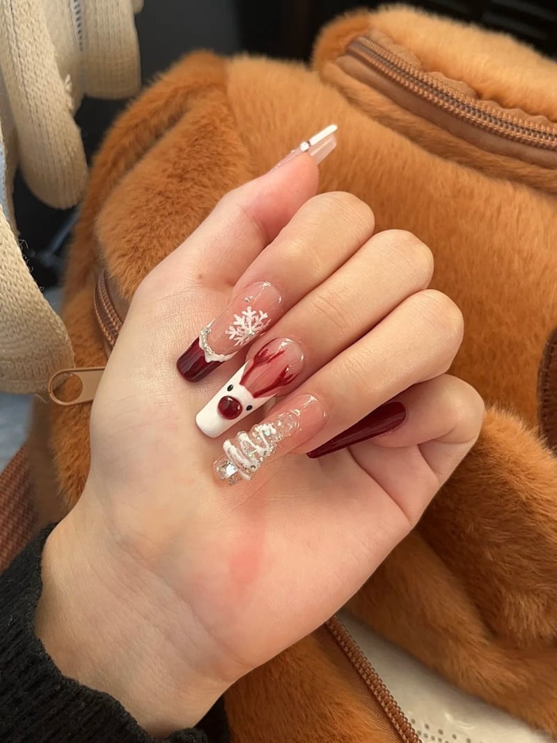 Bàn tay với móng tay trang trí hình tuần lộc, bông tuyết và cây thông, nền là túi xách lông màu nâu.