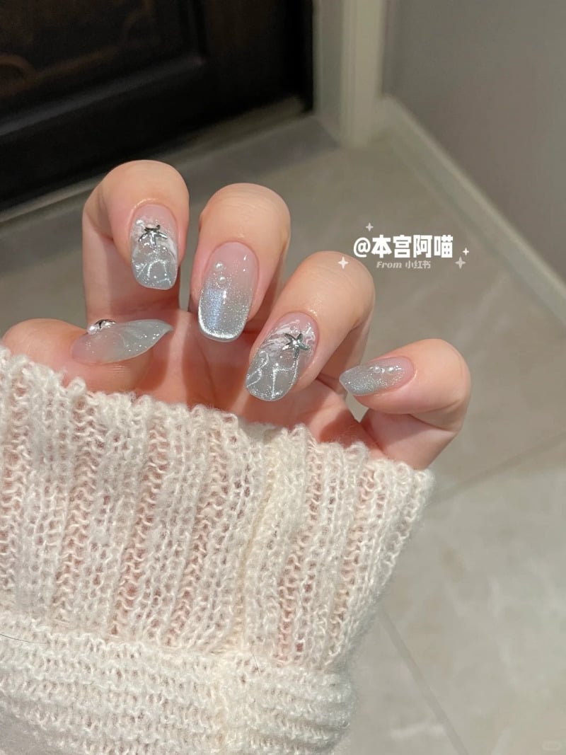 Tay đeo áo len trắng, móng tay được sơn màu bạc lấp lánh với họa tiết trang trí hình ngôi sao và hạt nhỏ.