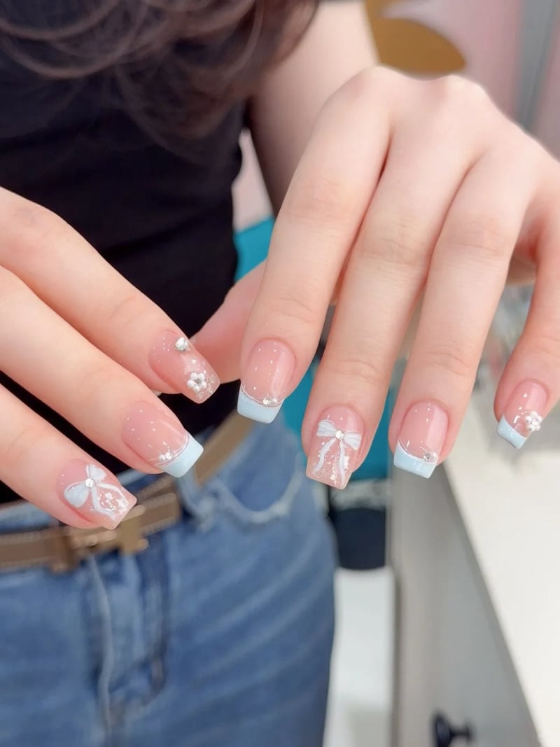 Bàn tay với móng tay được trang trí họa tiết nơ và hoa trắng, kiểu Pháp, trên nền quần jeans xanh.