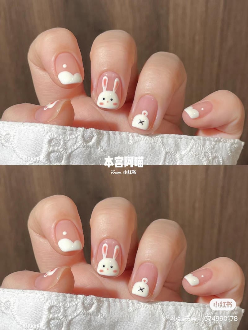 Bàn tay với móng tay trang trí hình thỏ và mây, nền màu hồng nhạt, có chi tiết trắng nổi bật.