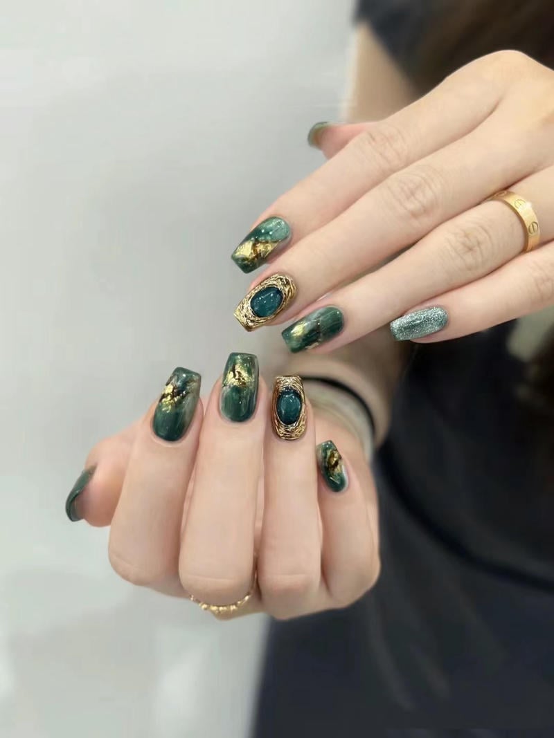Bàn tay với móng tay được sơn màu xanh lá cây, trang trí họa tiết vàng và đá xanh, đeo nhẫn vàng.