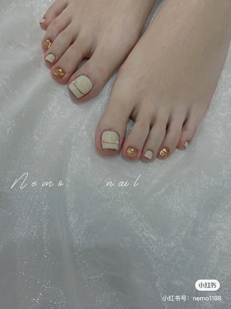 Bàn chân với móng được sơn màu be, trang trí họa tiết vàng lấp lánh, trên nền vải trắng.