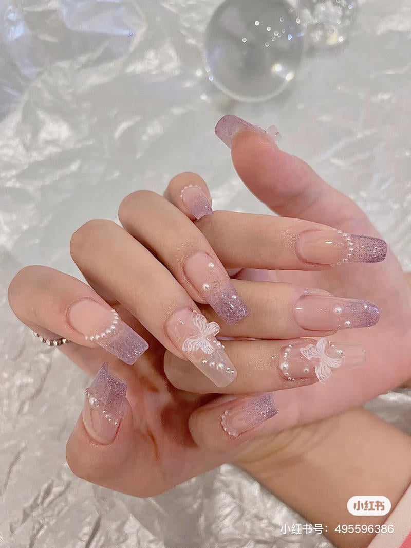Bàn tay với móng tay được trang trí màu tím nhạt, đính hoa văn và hạt cườm nhỏ trên nền vải trắng.