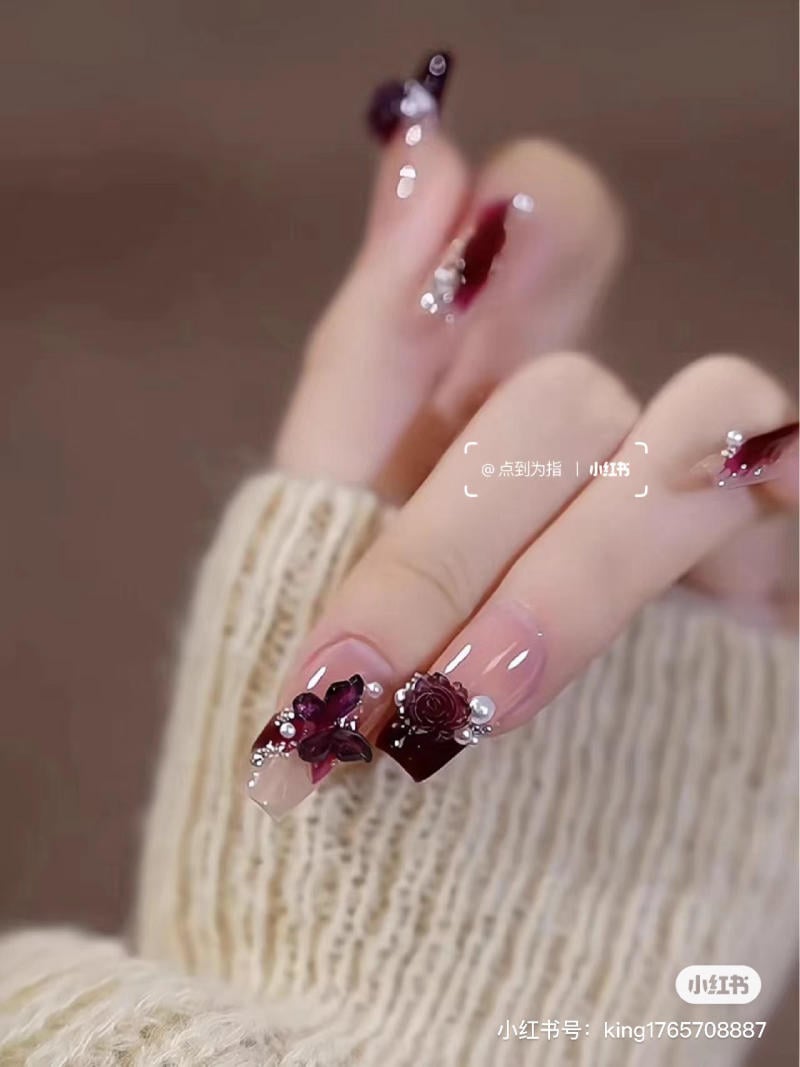 Bàn tay với móng tay được trang trí hoa và ngọc trai trên nền màu đỏ và trong suốt, mặc áo len màu kem.