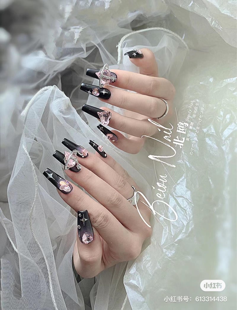 Bàn tay với móng tay màu đen được trang trí bằng đá và hoa văn, đặt trên nền vải trắng mềm mại.