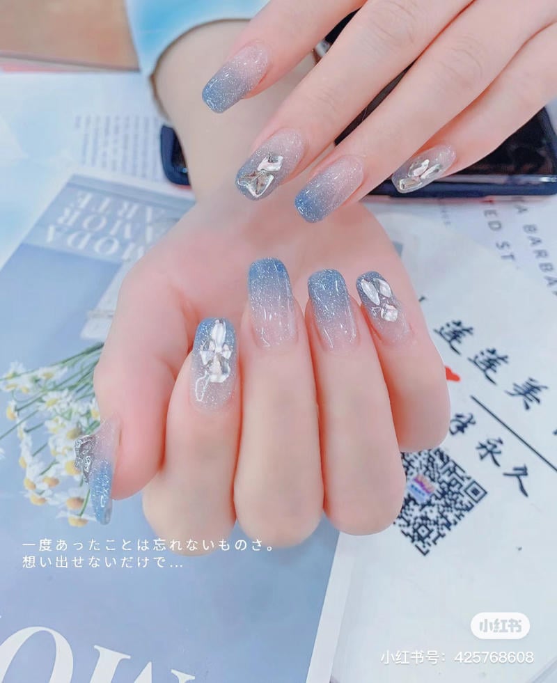 Bàn tay với móng tay được trang trí ombre xanh và bạc, có đính đá lấp lánh, đặt trên tạp chí.