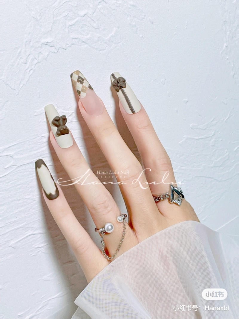 Bàn tay với móng tay dài được trang trí họa tiết nơ và caro, đeo nhiều nhẫn bạc có hạt ngọc trai.