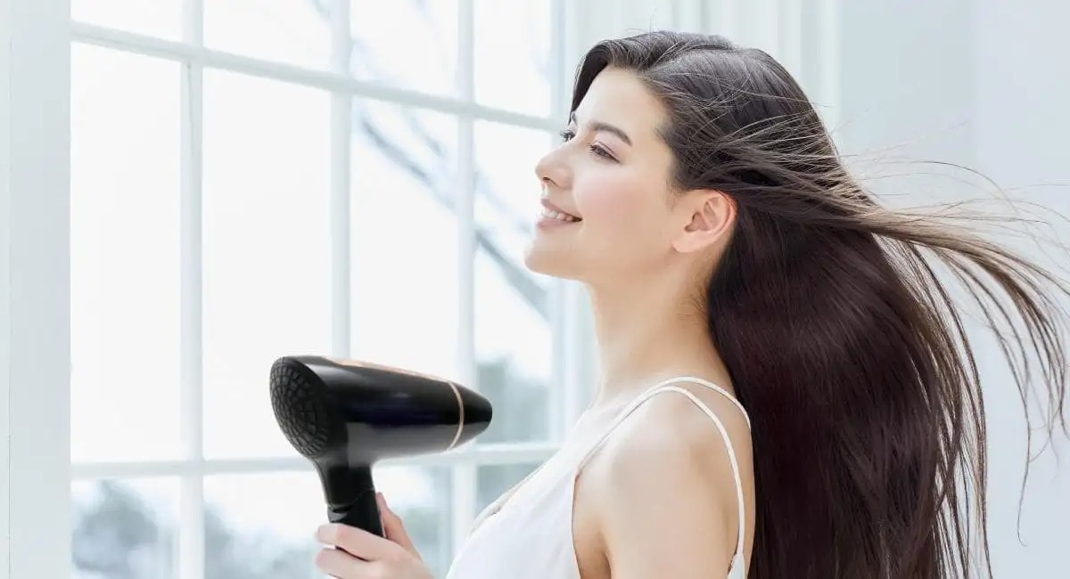Người cầm máy sấy tóc, gió thổi qua mái tóc dài, đứng cạnh cửa sổ sáng.