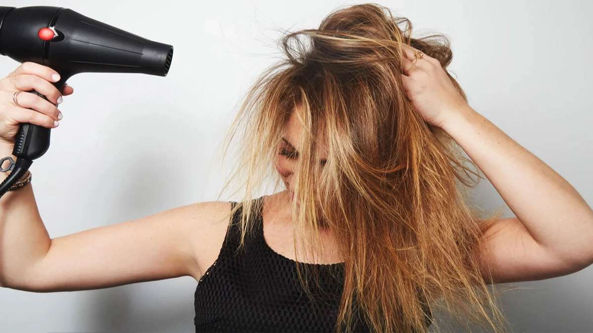 Người đang sấy tóc với máy sấy đen, tay còn lại giữ tóc, nền trắng.