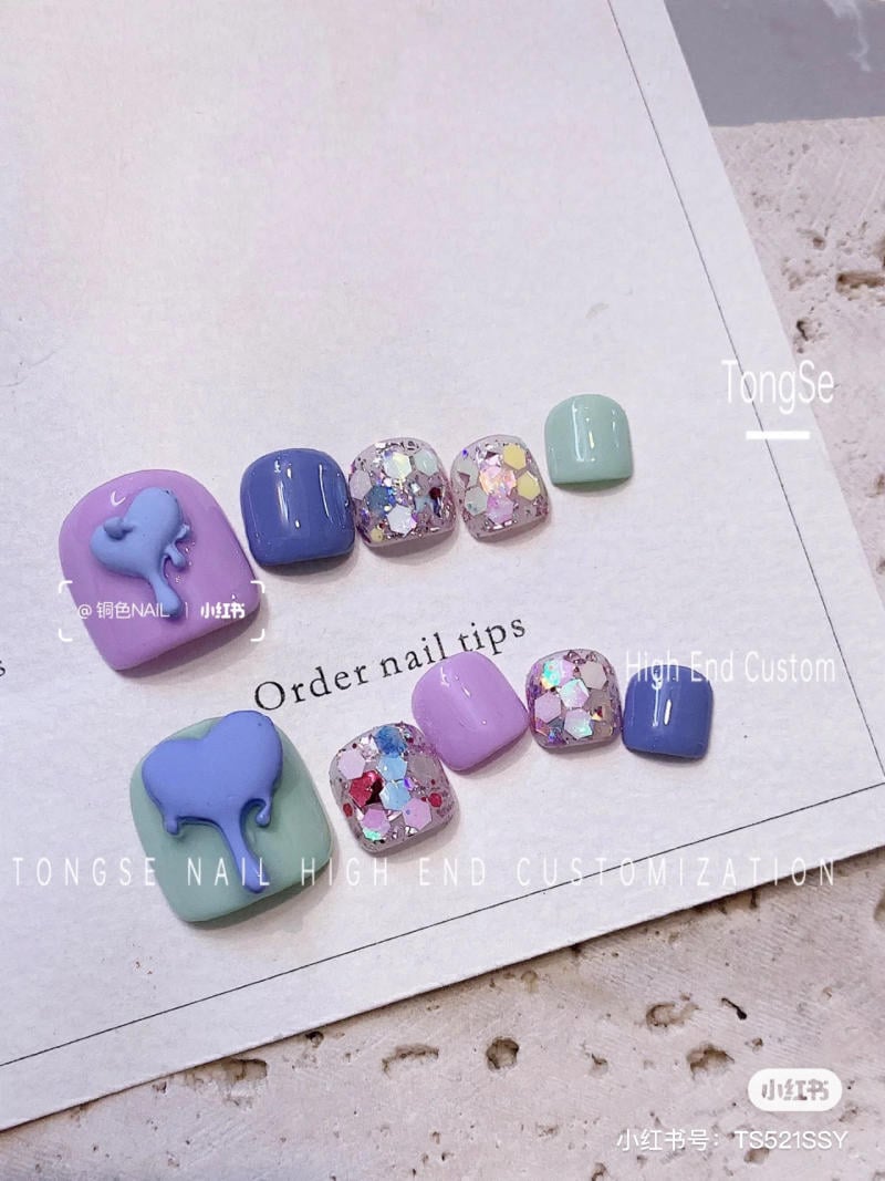 Bộ móng giả với họa tiết tim xanh và kim tuyến nhiều màu sắc trên nền giấy.