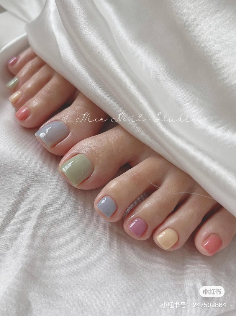 Bàn chân với móng được sơn nhiều màu pastel, đặt trên nền vải trắng.
