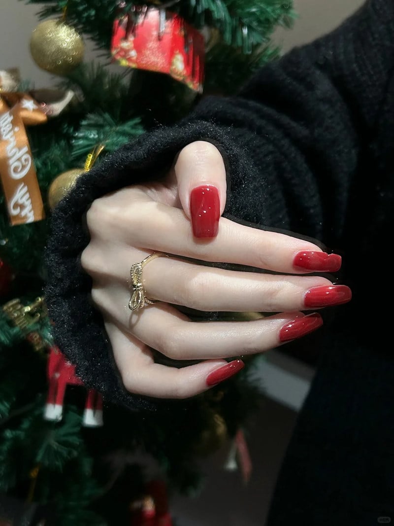 Tay với móng sơn đỏ, đeo nhẫn, phía sau là cây thông Noel trang trí lấp lánh.