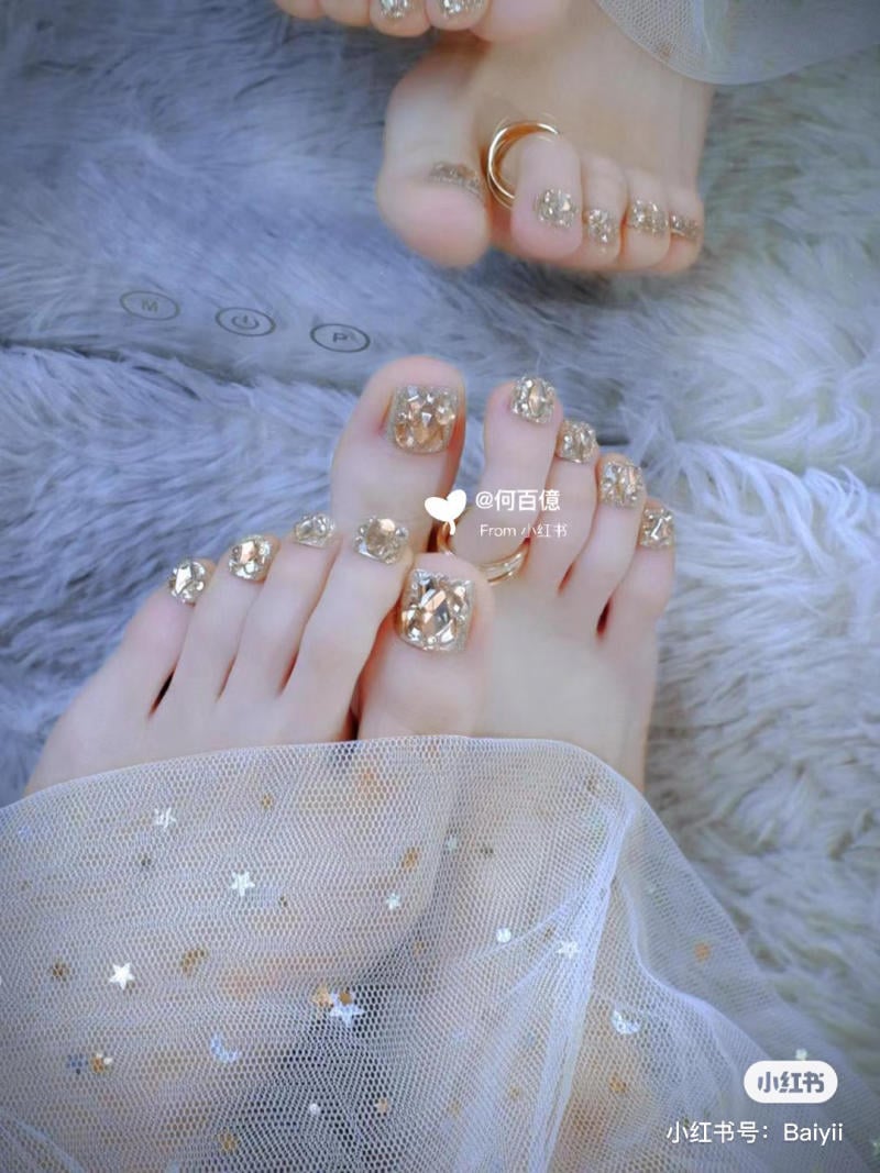 nai-chan-dinh-da__13__3204b7d7f732463ebfc5f9fcfe555102 Bàn chân với móng được trang trí họa tiết lấp lánh, đeo nhẫn, đặt trên nền lông mềm và vải lưới có hoa văn sao.