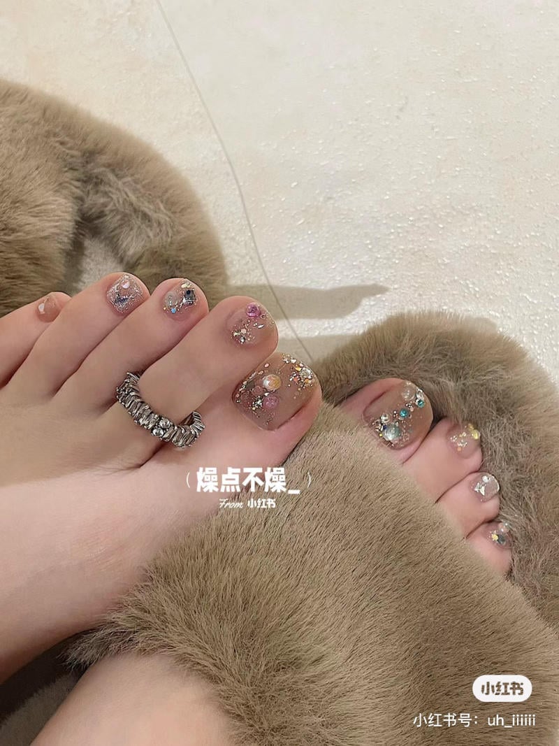nai-chan-dinh-da__3__5fd307868acc444794eccf88c962ad43 Bàn chân với móng được trang trí lấp lánh, đeo nhẫn ngón chân, trên nền dép lông màu nâu.