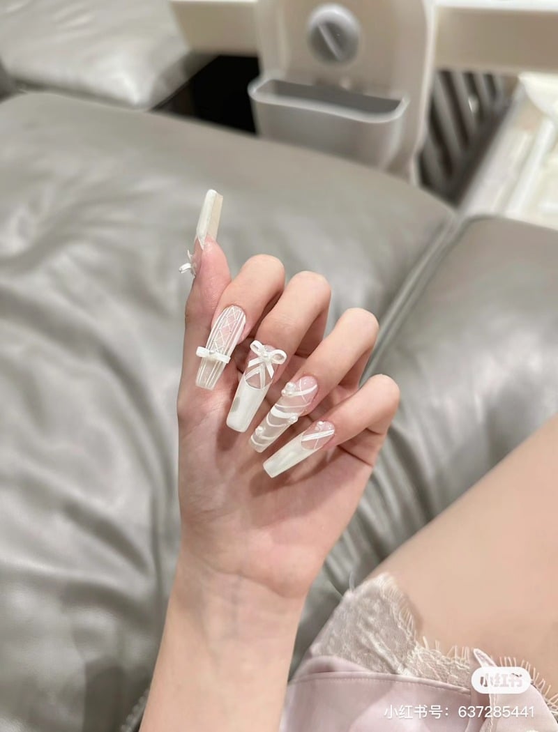 Bàn tay với móng tay dài được trang trí họa tiết trắng tinh xảo, đặt trên nền ghế sofa màu xám.