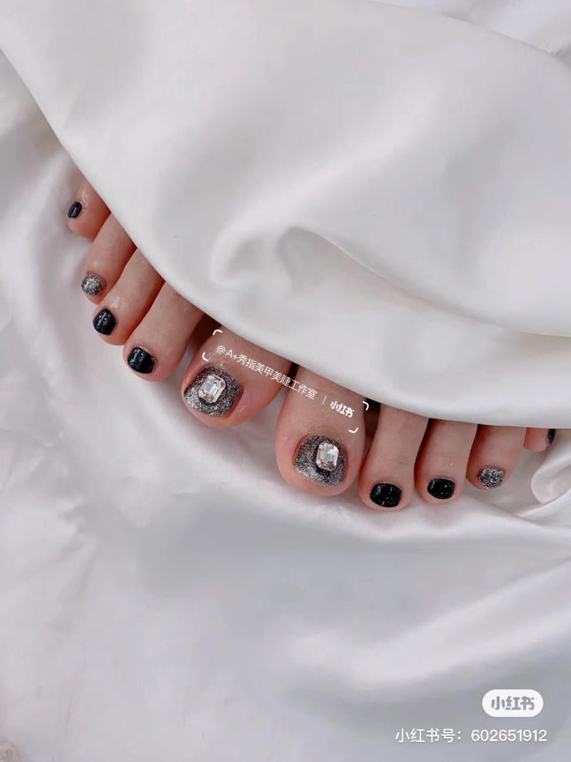 nail-chan-dinh-da__10__b10b43c6acbd4d08ad2b9f4262d7d7df Bàn chân với móng được sơn màu đen và bạc lấp lánh, đặt trên nền vải trắng.