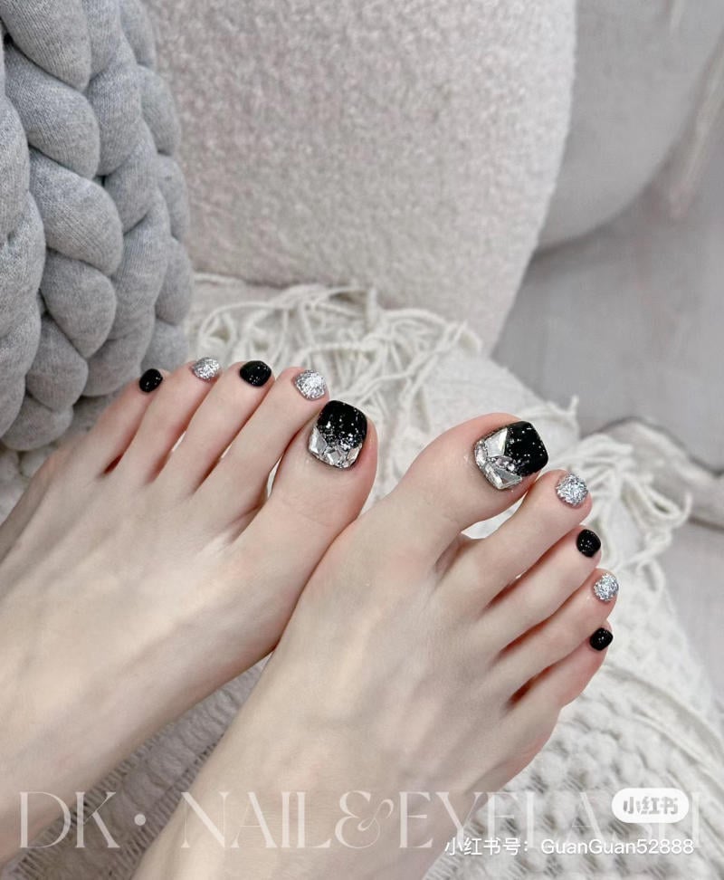 nail-chan-dinh-da__13__fa74c74f7b1c470a88d6702395a87b14 Bàn chân với móng chân sơn màu đen và bạc, đặt trên nền vải mềm màu xám nhạt.