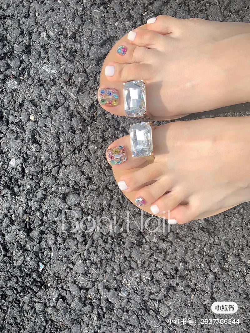 nail-chan-dinh-da__16__f6d84a2b728d4328baf4314b4af68831 Bàn chân với móng được trang trí họa tiết đa sắc, đeo dép có chi tiết đá lớn, đứng trên nền đường nhựa.