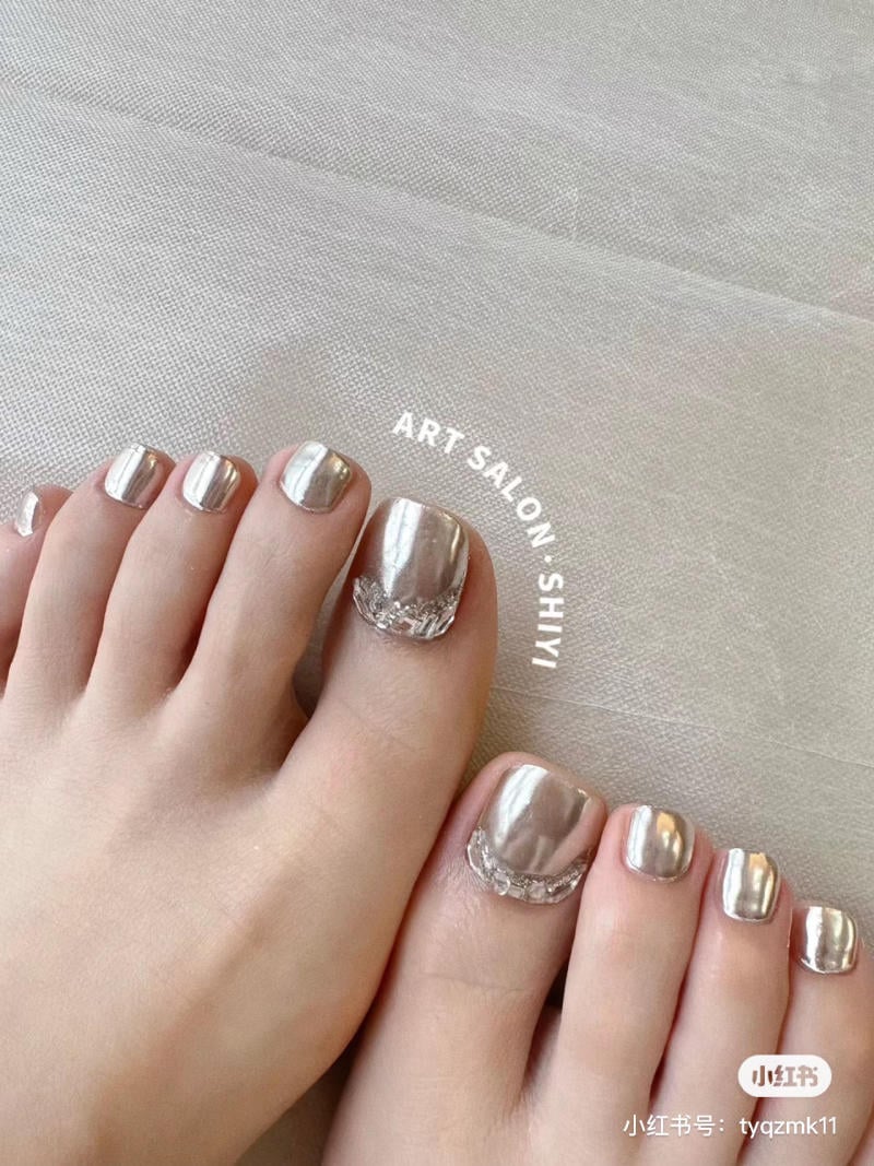 nail-chan-dinh-da__1__34bd7c0000014d31b3d3697f13b9eb5b Bàn chân với móng được sơn màu bạc ánh kim, nằm trên bề mặt vải màu nhạt.