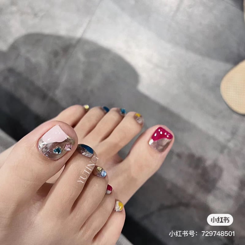 nail-chan-dinh-da__20__729e139c1f39406ba907cbe0e02aee30 Bàn chân với móng chân được sơn nhiều màu sắc và trang trí đá lấp lánh trên nền sàn xám.