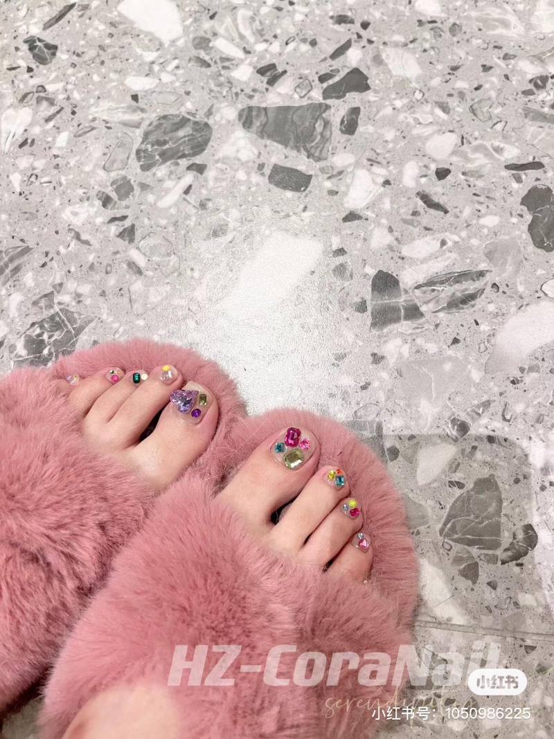 nail-chan-dinh-da__21__d8f45479c02a4a57aa6963f87315cf7c Bàn chân với móng chân được trang trí nhiều màu sắc, mang dép lông màu hồng trên sàn đá hoa văn xám.