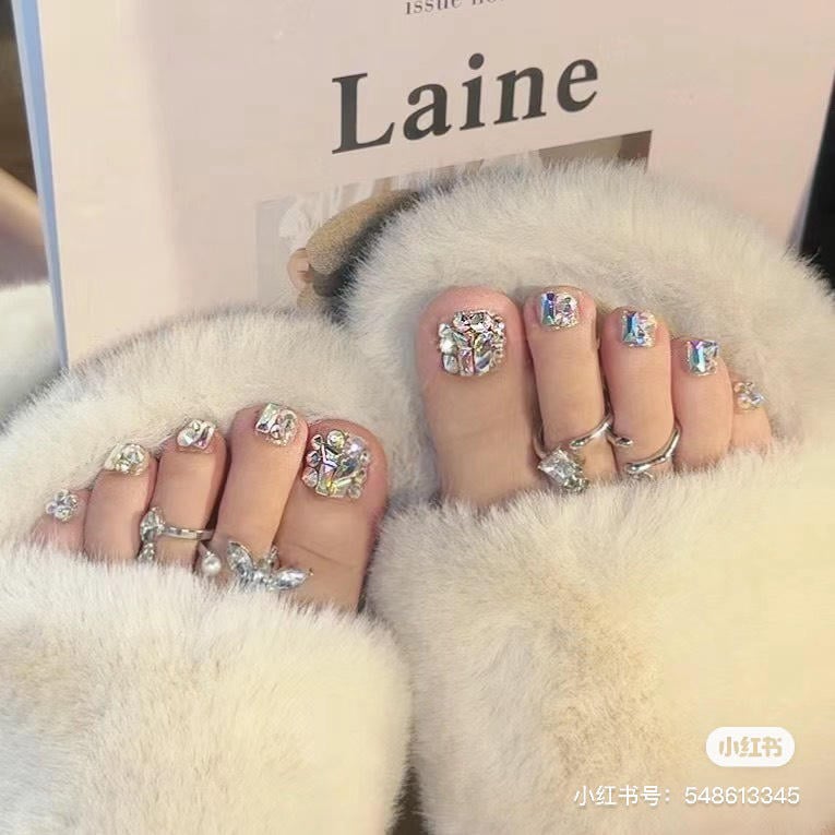 nail-chan-dinh-da__22__526dfe4d9f92409f8568388e45c3a57d Đôi chân với móng chân trang trí lấp lánh, đeo nhẫn, đặt trên dép lông, bên cạnh cuốn sách.