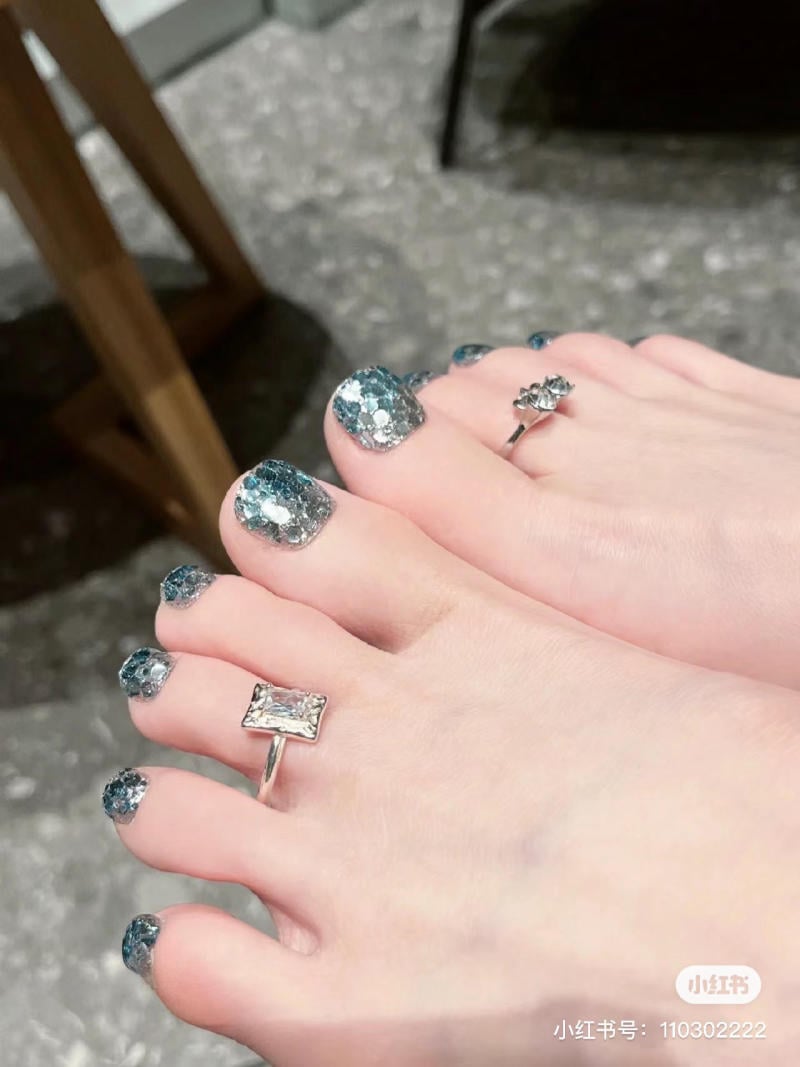 nail-chan-dinh-da__24__fadcadf0e8bb42a7a01f1342b998b359 Bàn chân với móng chân sơn kim tuyến xanh và hai chiếc nhẫn trang trí, đặt trên sàn gạch xám.