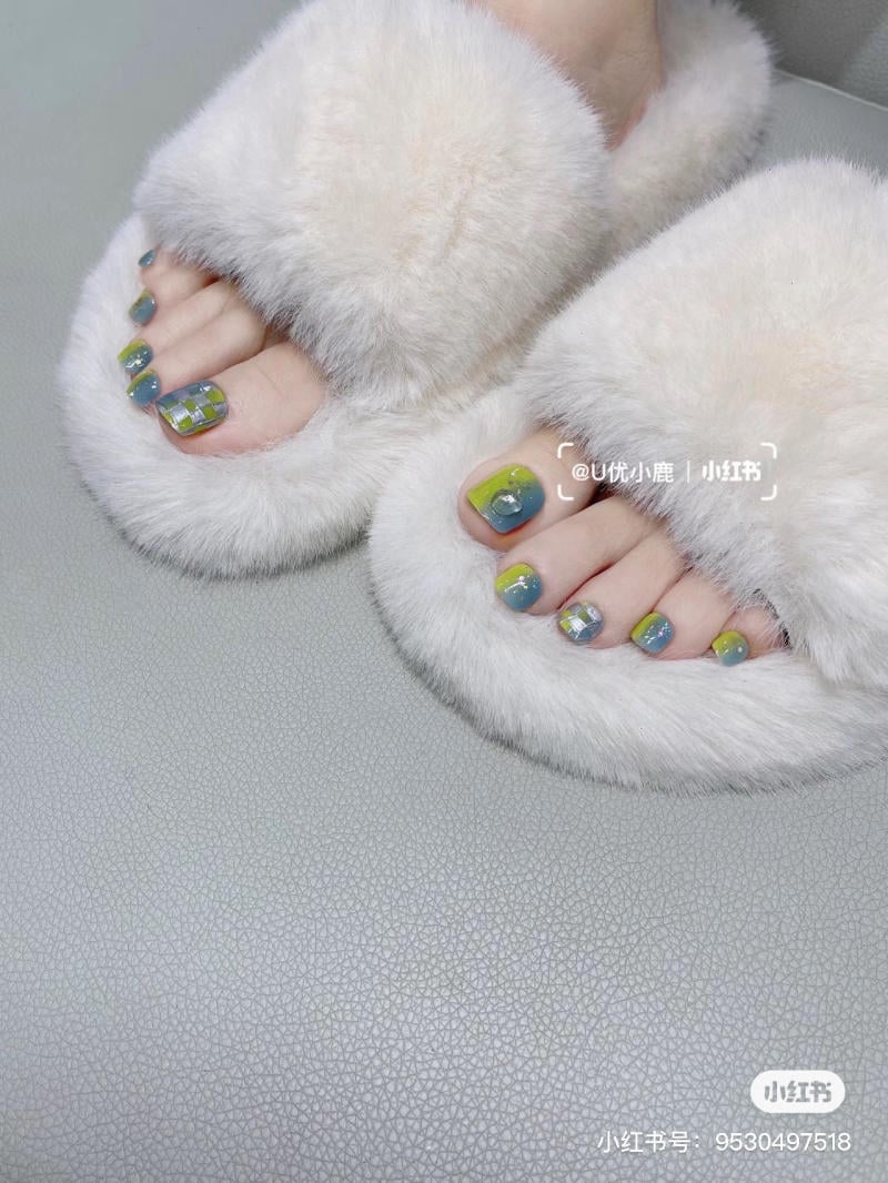 nail-chan-dinh-da__37__bbb0592e3a894b4db3da7b62ea7f37bd Bàn chân với móng được sơn màu sắc và họa tiết, đi dép lông màu trắng trên bề mặt màu xám.
