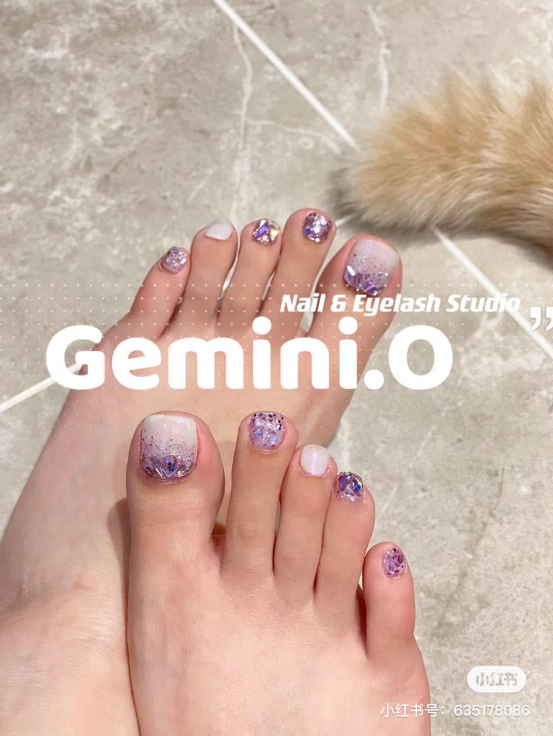 nail-chan-dinh-da__38__2ac8f8b72fe7430eac70f06eb6befe99 Bàn chân với móng được trang trí họa tiết màu tím và trắng trên nền sàn gạch xám.