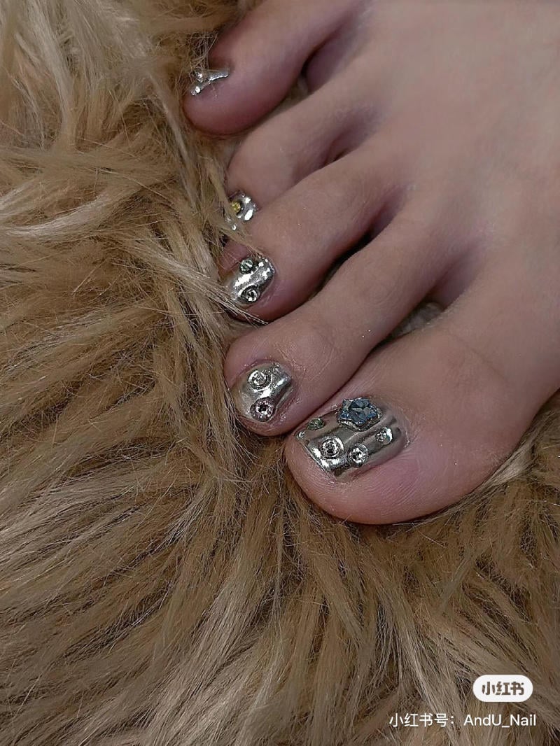 nail-chan-dinh-da__3__f6764dc5395a4b7b83510080c73cbda0 Bàn chân với móng chân được sơn màu bạc, trang trí đá lấp lánh, đặt trên bề mặt lông màu nâu nhạt.
