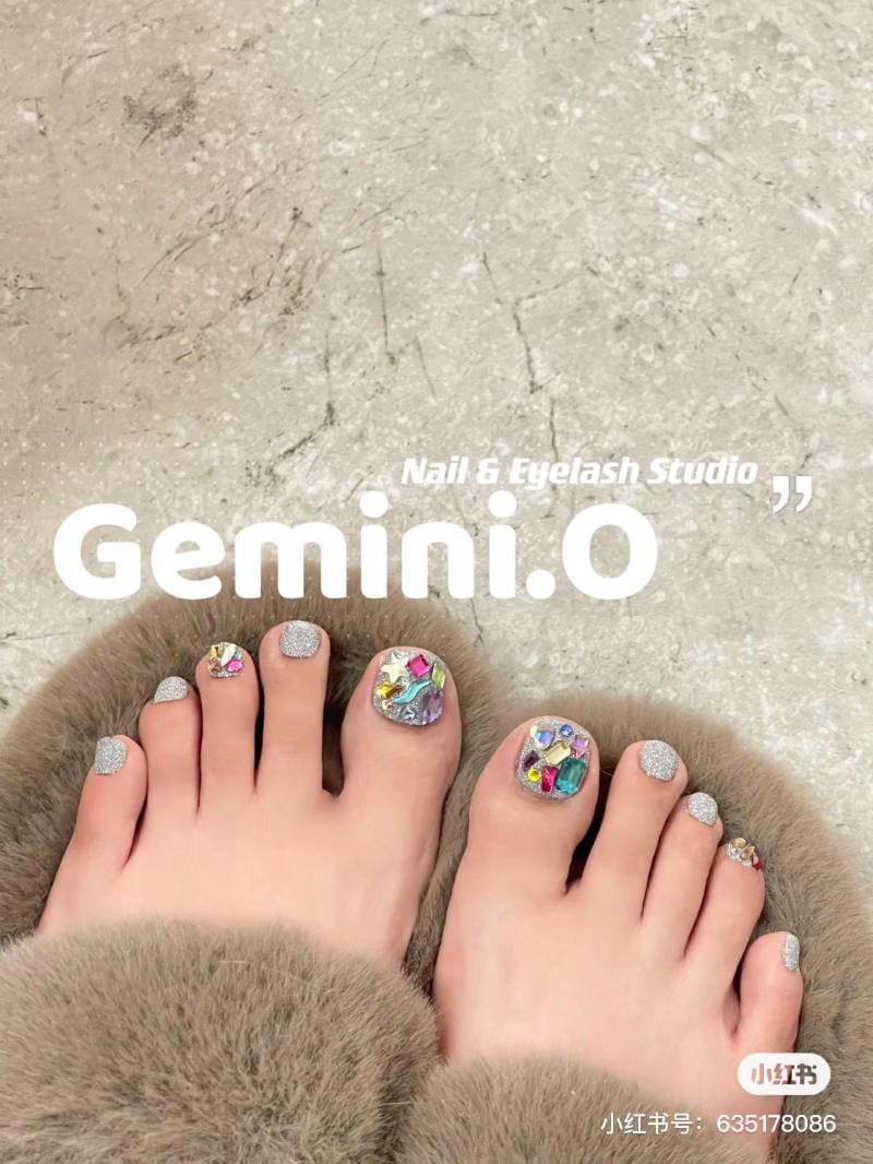 nail-chan-dinh-da__46__3ab56daf92fa48ef80d9607218ec8c49 Bàn chân với móng được trang trí đá quý và kim tuyến, đặt trên bề mặt lông mềm, có chữ "Gemini.O".