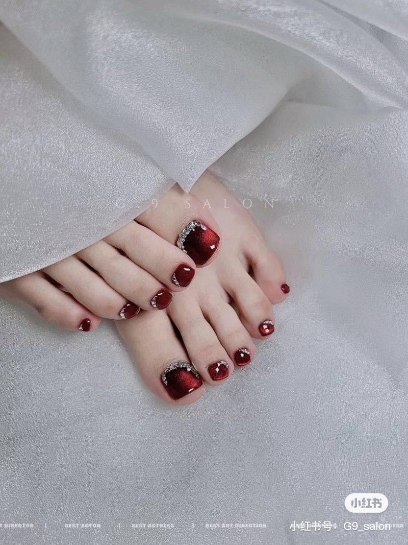 nail-chan-dinh-da__51__522d4547f8bc4017984947d8911cc357 Bàn chân với móng được sơn màu đỏ bóng, trang trí đá quý, trên nền vải trắng sáng bóng.