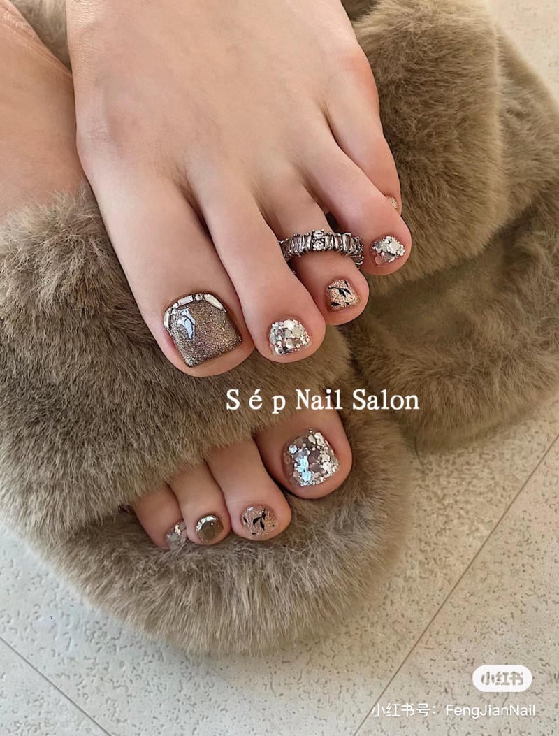 nail-chan-dinh-da__53__211768f91a63437298a1219148ff9c83 Ngón chân với móng được sơn kim tuyến và đeo nhẫn, mang dép lông màu nâu trên sàn nhà.