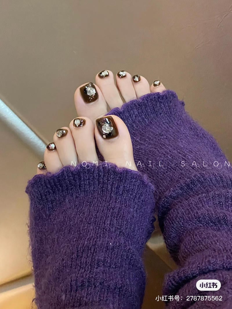 nail-chan-dinh-da__54__0a33595039f34dddb21ace64d4061188 Bàn chân với móng chân sơn màu tối, gắn đá trang trí lấp lánh, mang tất màu tím.