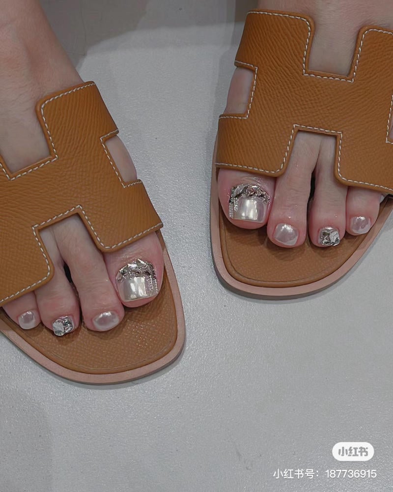 nail-chan-dinh-da__5__e91a745ce9624b3281ff1a6f41d77751 Đôi chân mang dép nâu, móng chân được sơn màu bạc lấp lánh với các chi tiết trang trí.