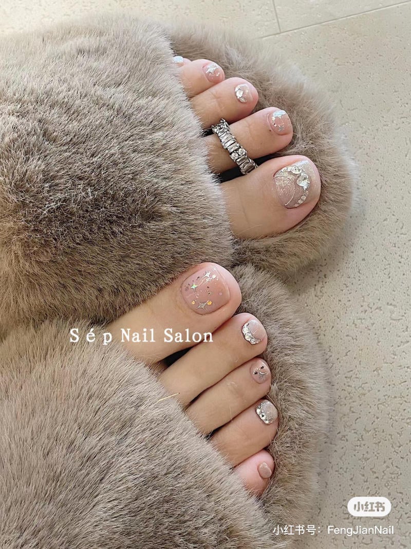 nail-chan-dinh-da__6__e3d76fb3a9084f1e9d2cfa7233fb2736 Bàn chân với móng chân được trang trí họa tiết lấp lánh, đi dép lông màu xám nhạt, có nhẫn ở ngón chân.