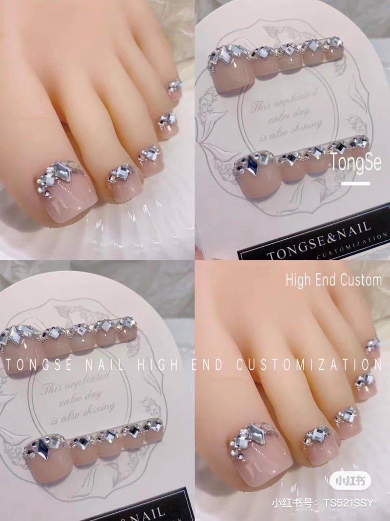 nail-chan-dinh-da__8__6d5a393530e24f899a0b855e80efa450 Mẫu móng chân giả đính đá lấp lánh, nền hồng nhạt, được trưng bày trên giá đỡ trang trí hoa văn.