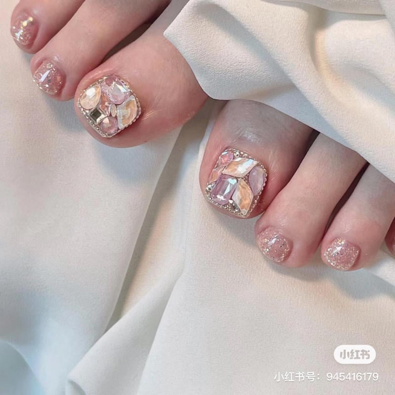 nail-chan-dinh-da__9__cbd53ead44fa4dd991c57421cf9d7712 Bàn chân với móng được trang trí lấp lánh và đính đá, trên nền vải trắng mềm mại.