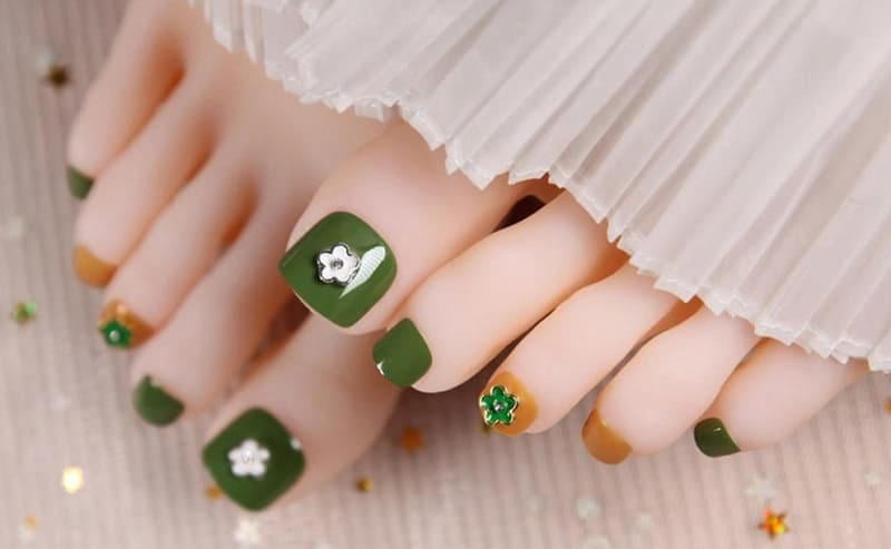 Bộ móng chân được sơn màu xanh lá cây với họa tiết hoa trang trí, nền vải màu trắng xếp ly.