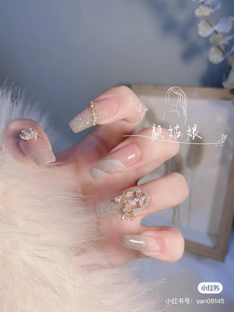 Bàn tay với móng tay giả màu lấp lánh, trang trí đá quý và hoa văn tinh xảo, phông nền mờ.