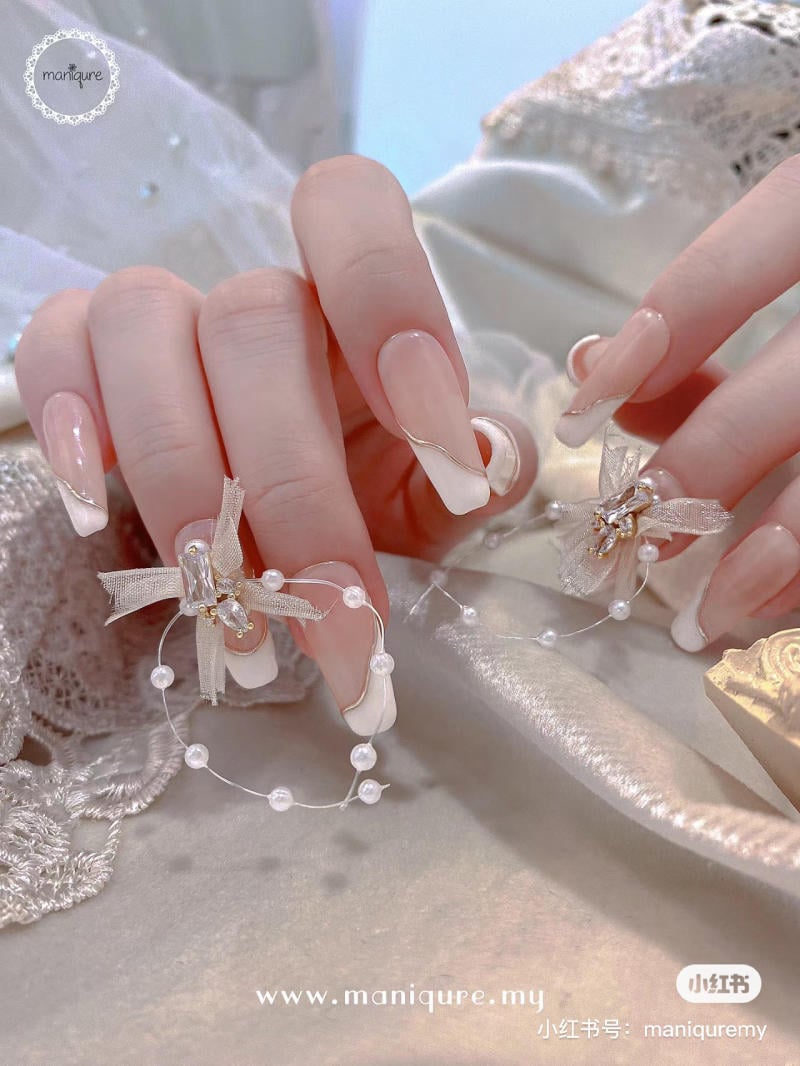 Bàn tay với móng được làm kiểu Pháp, trang trí bằng vòng ngọc trai và nơ trên nền vải trắng.