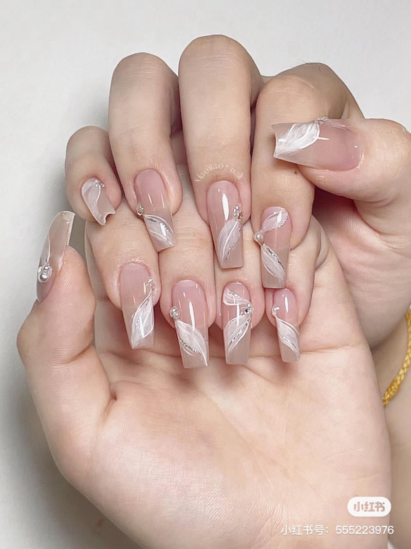 Bàn tay với móng tay dài được trang trí họa tiết trắng và đá lấp lánh trên nền màu nude bóng.