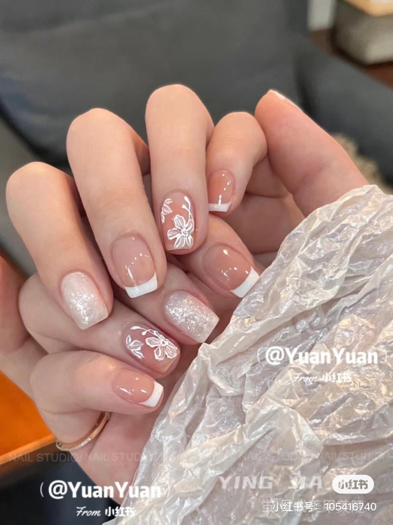 Bàn tay với móng tay sơn màu nude, họa tiết hoa trắng và viền trắng, cầm một mảnh giấy bóng.