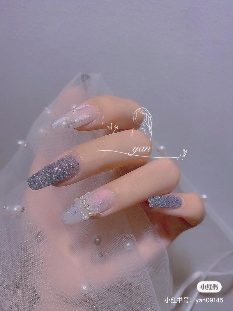 Bàn tay với móng tay được sơn màu xám và trắng, trang trí hạt ngọc trai và nơ, trên nền vải lưới.